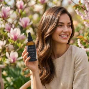 Усмихната жена държи кокосово масло за коса сред цъфнали магнолии в слънчев пролетен ден.