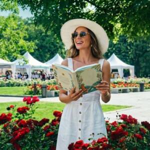 📚 Панаир на книгата 2026: Моите книжни находки, топли заглавия и летни изкушения Засмяна жена с бяла шапка чете книга пред шатрите на Панаир на книгата 2026 сред рози.